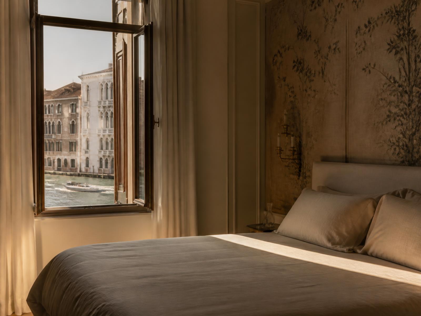 Palazzo bedroom in oatmeal linen beside an open window looking onto the Grand Canal at golden hour