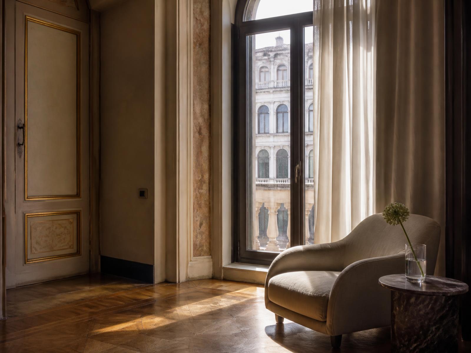 Corner of a Venetian suite with a pale linen armchair, sheer curtain and parquet floor in late afternoon light