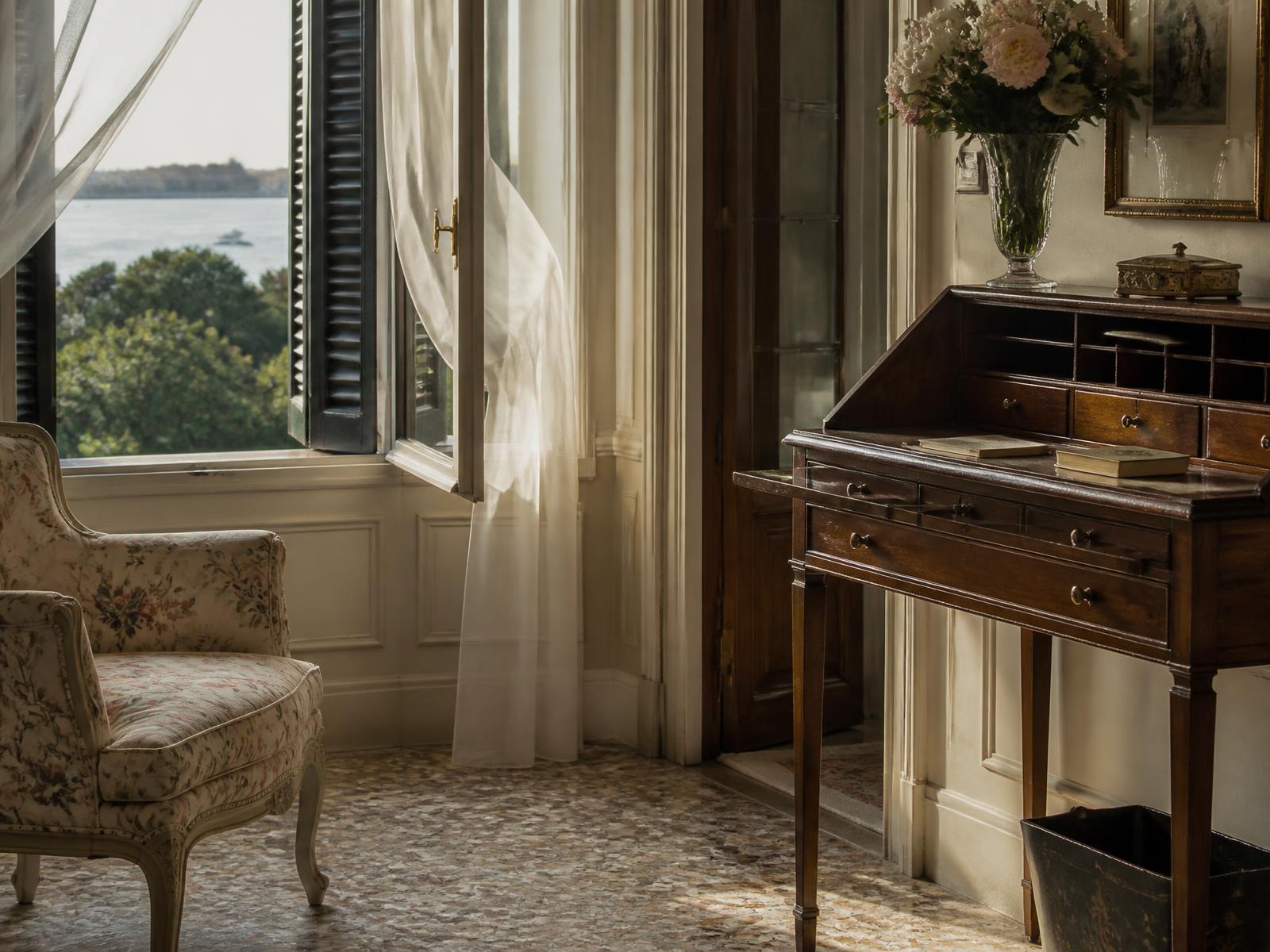 Corner of a Venetian suite with a hand-painted floral armchair, antique writing desk and sheer curtain lifting at an open window