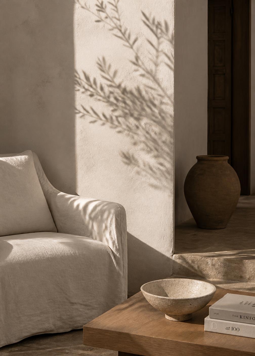 Corner of a Mediterranean suite with linen armchair, handmade ceramic bowl, plaster wall and the shadow of an olive branch in afternoon light