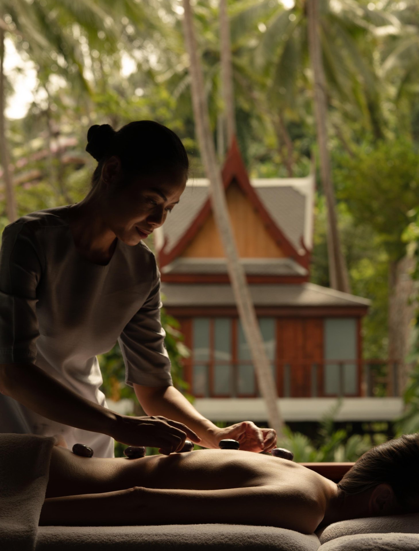 Behandlung mit warmen Steinen in einem Raum des Aman Spa, dahinter durch das Fenster ein thailändischer Pavillon