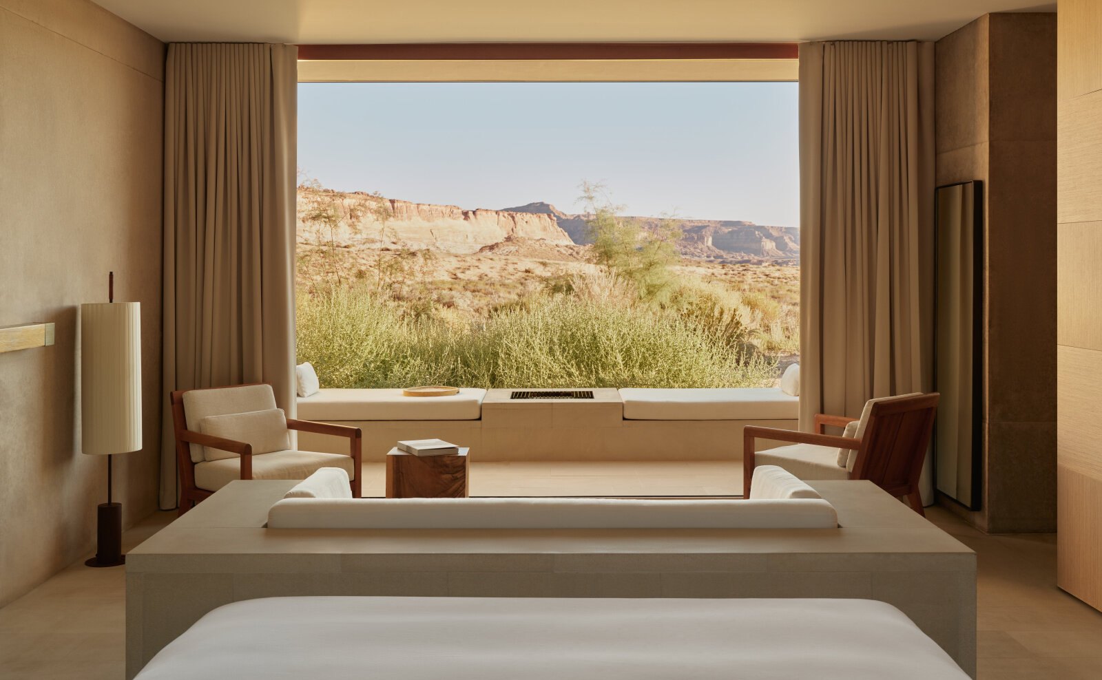 Desert Suite in Amangiri: niedriges Bett vor einem raumhohen Fenster, das die Wüste wie ein Bild rahmt, davor zwei Sessel aus hellem Holz und eine Fensterbank mit Kamin