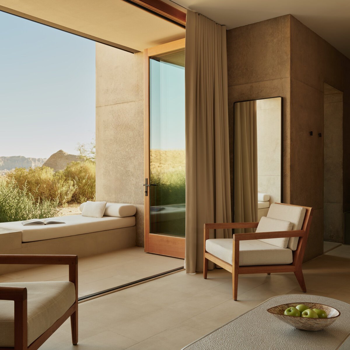 Wohnbereich einer Desert Suite mit gepolsterter Fensterbank, offener Tür zur Terrasse, Standspiegel und Sesseln aus Kirschholz im Vormittagslicht