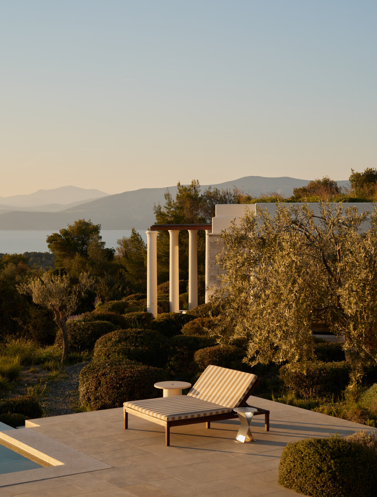 Terrasse in Amanzoe im Abendlicht: gestreifte Liege am Pool, dahinter drei helle Säulen zwischen Olivenbäumen und die Hügel über dem Golf