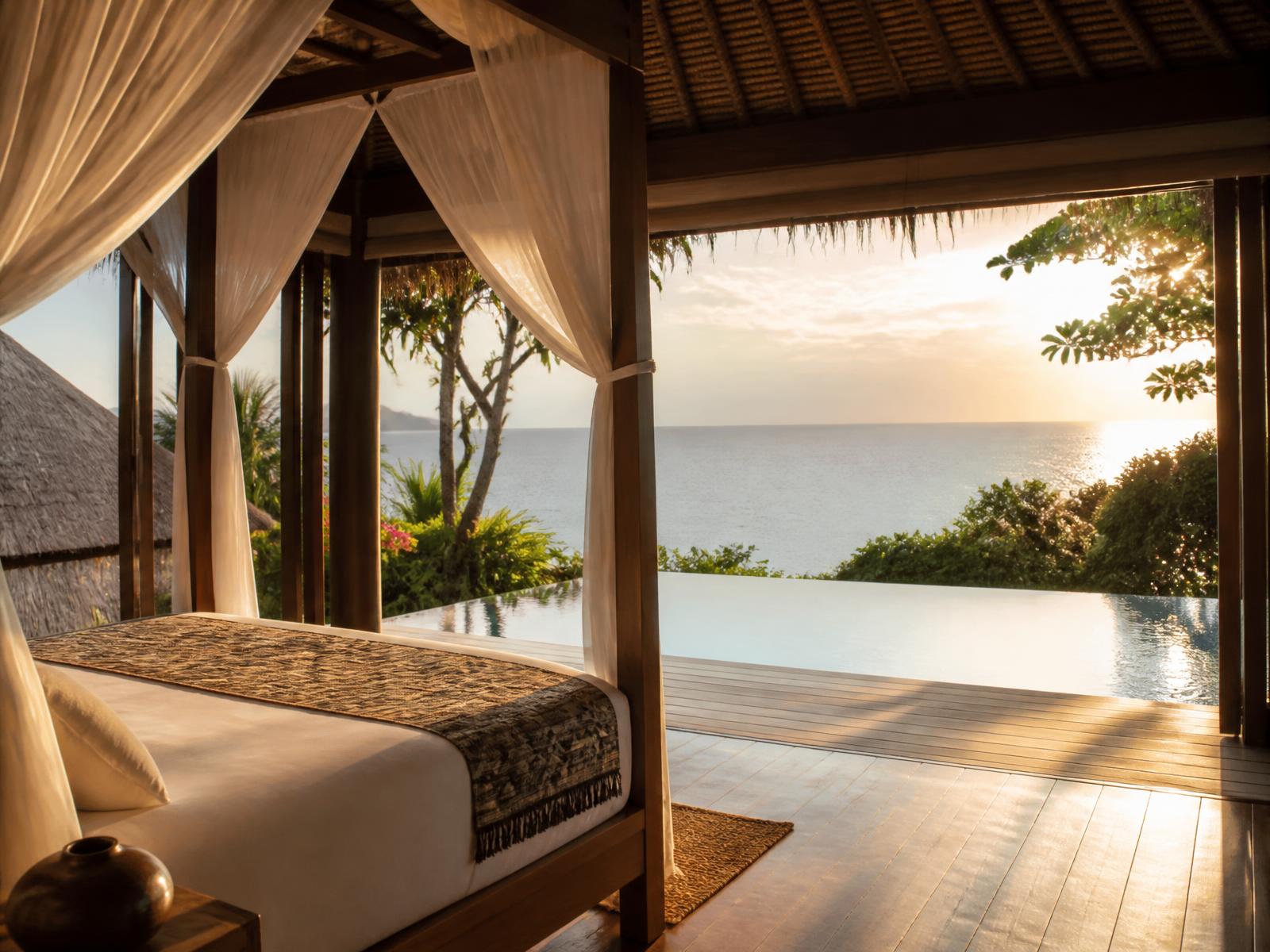 Open-air Indonesian villa bedroom with teak four-poster bed, white linen, thatched ceiling and no fourth wall, opening onto a private pool and the Indian Ocean at sunrise