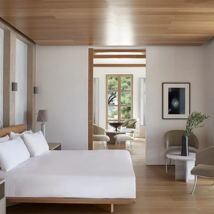Schlafzimmer einer Amanzoe-Villa mit heller Holzdecke, weißem Bett und offener Verbindung zu einem Sitzraum mit Sprossenfenster