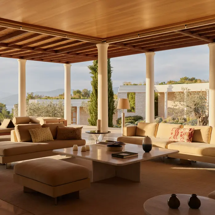 Offener Wohnbereich einer Amanzoe-Villa unter Holzdecke zwischen Säulen, mit tiefen Sofas, niedrigem Steintisch und Blick auf Zypressen, Olivenbäume und Meer