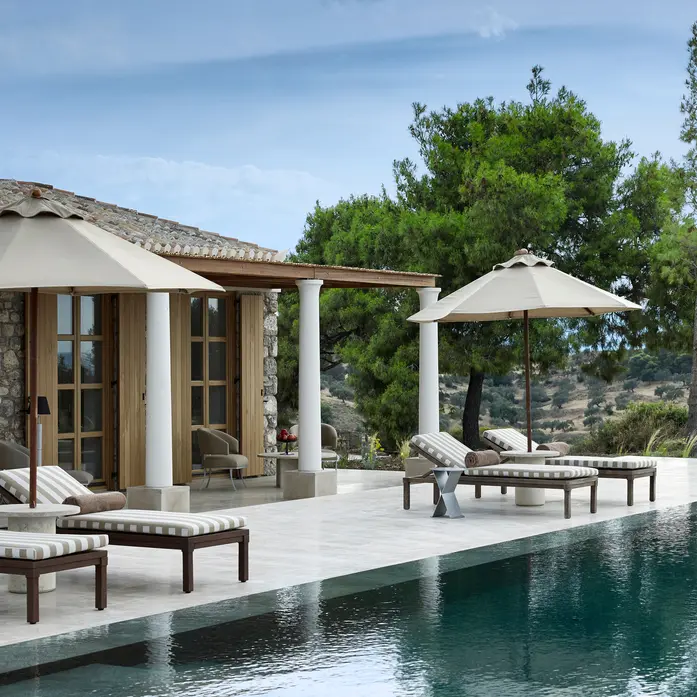 Privater Pool einer Amanzoe-Villa mit gestreiften Liegen, Sonnenschirmen, Natursteinmauer und Pinien über den Hügeln