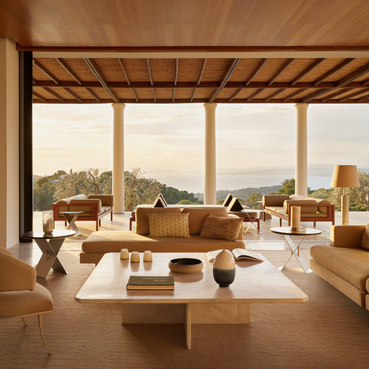 Wohnbereich einer Amanzoe-Villa im Abendlicht, offen zur Säulenterrasse mit Blick über Olivenbäume auf den Golf
