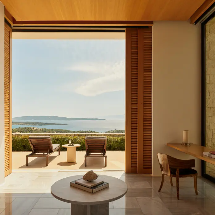 Blick aus einem Amanzoe-Zimmer durch geöffnete Holzflügel auf zwei Liegen, Hecken und den Golf mit Halbinsel