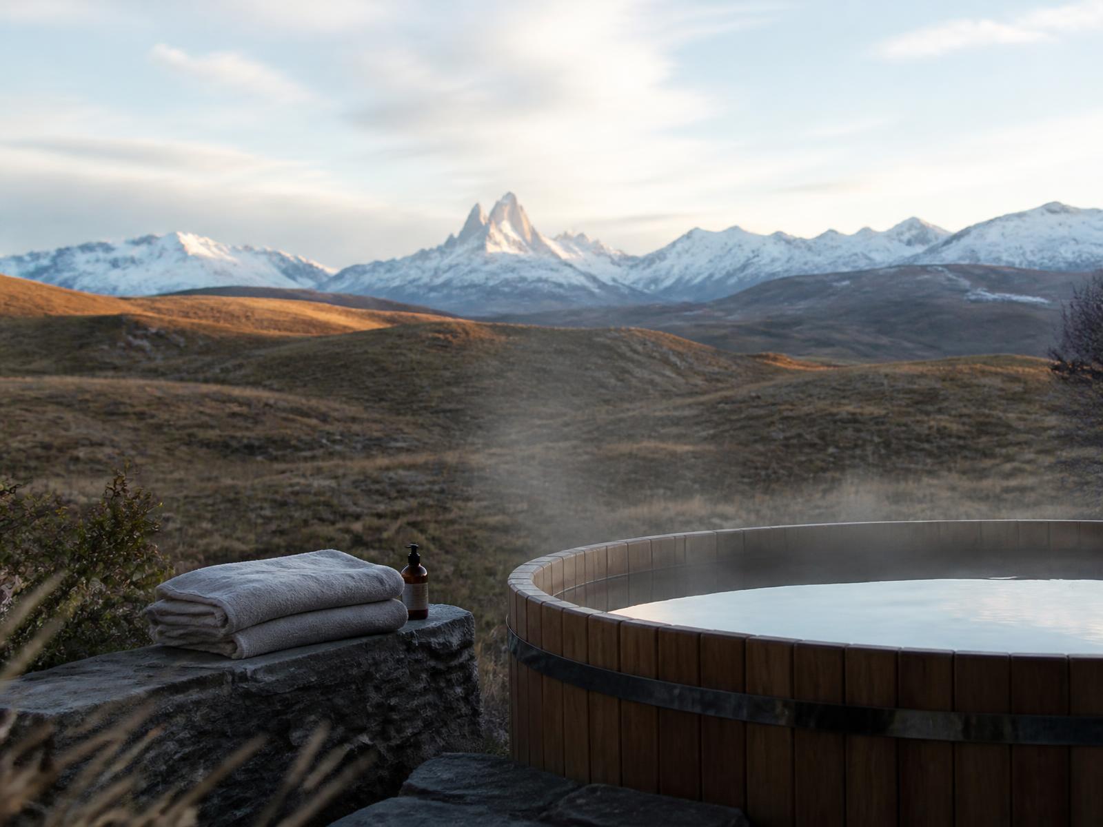 Outdoor timber hot tub steaming in cold Patagonian air, facing golden grassland and distant snow-covered peaks in late afternoon light