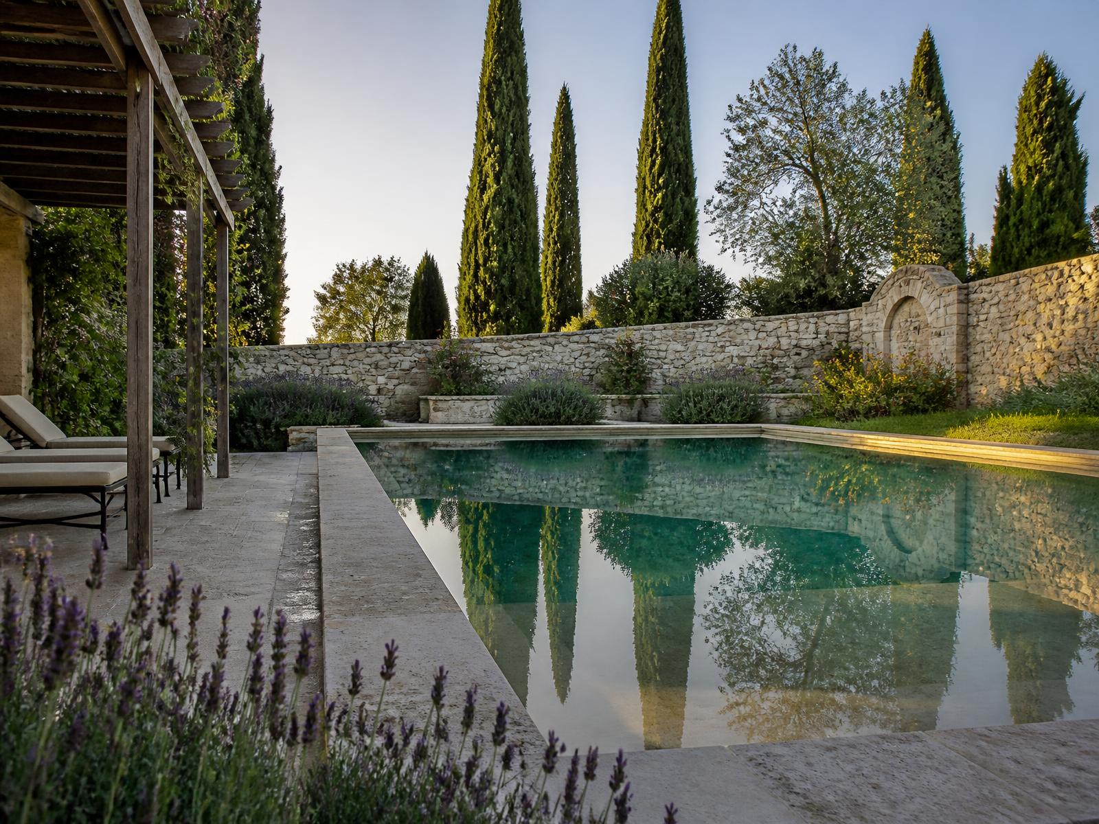 A still outdoor swimming pool in a private Italian garden with travertine surround, cypress trees, lavender and an old stone wall in golden afternoon light