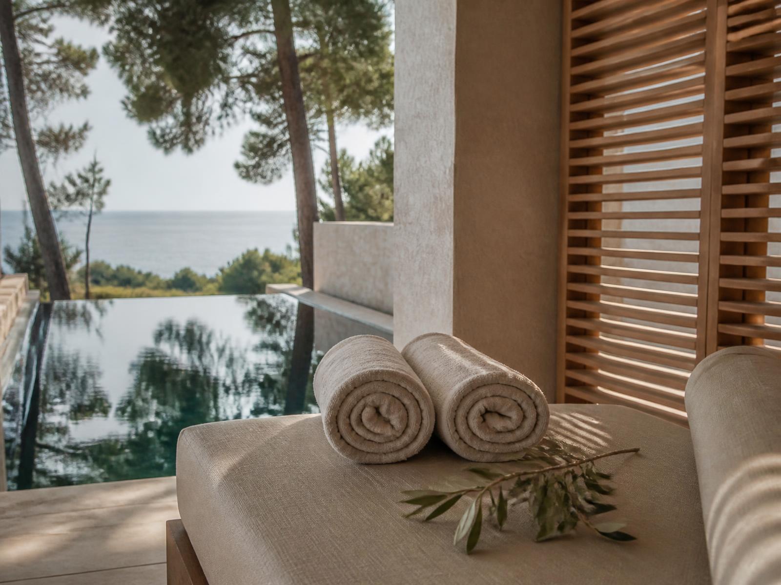 Quiet spa terrace with linen daybed, rolled towels and an olive branch beside a still pool reflecting pine trees