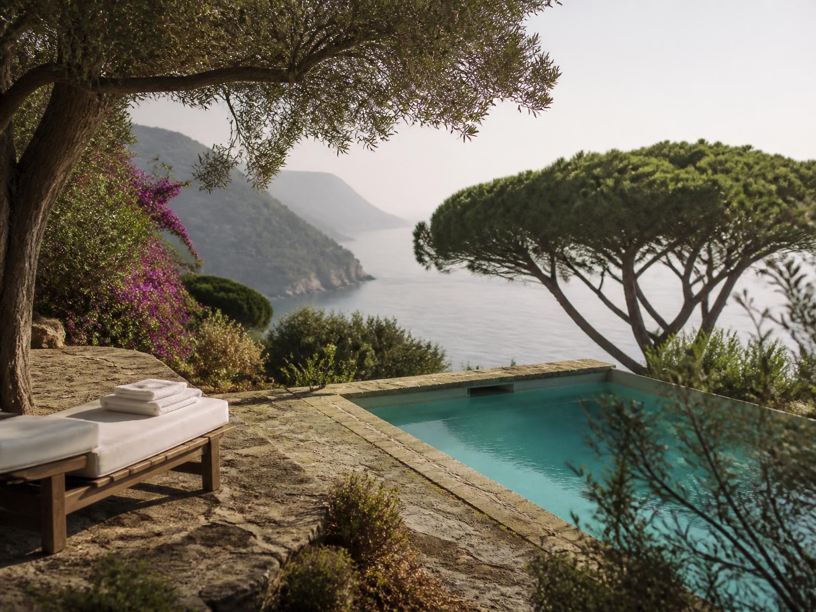 Quiet seawater swimming pool set into a terraced Mediterranean garden with olive trees, stone paving and the Ligurian coast beyond in soft morning light
