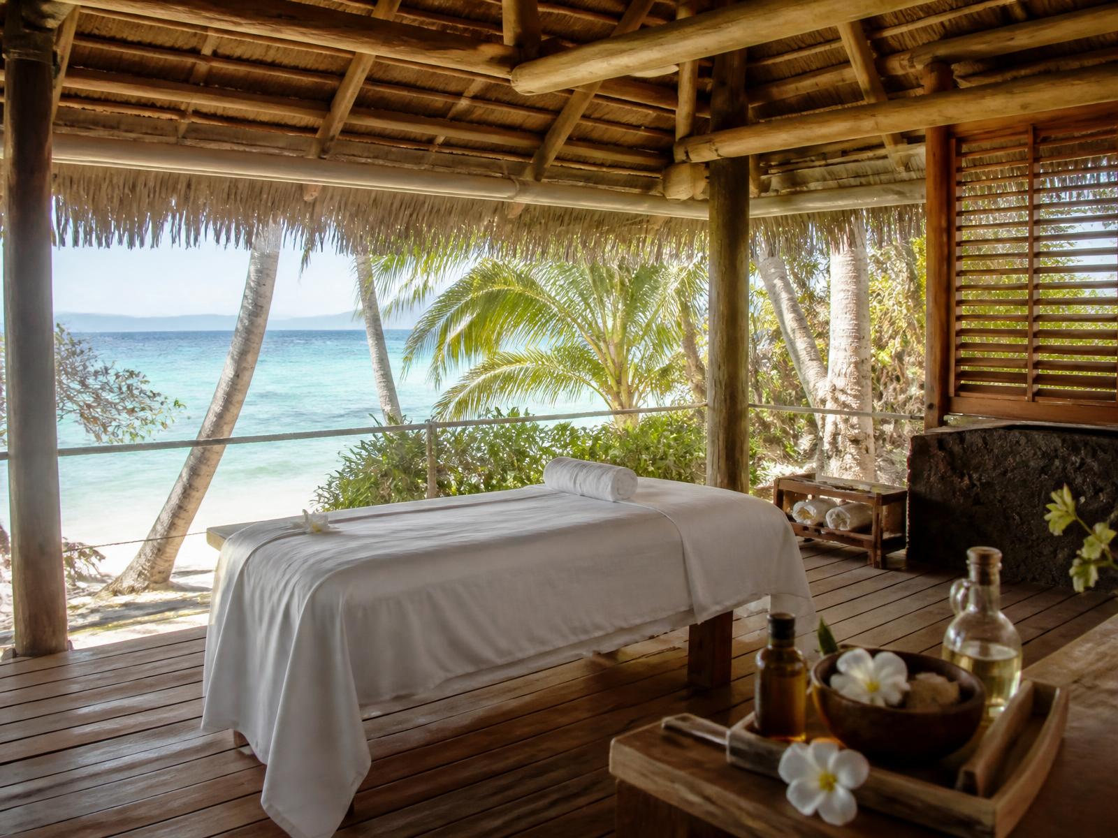 Open-air Polynesian spa pavilion with thatched roof and white linen among coconut palms, overlooking a turquoise lagoon