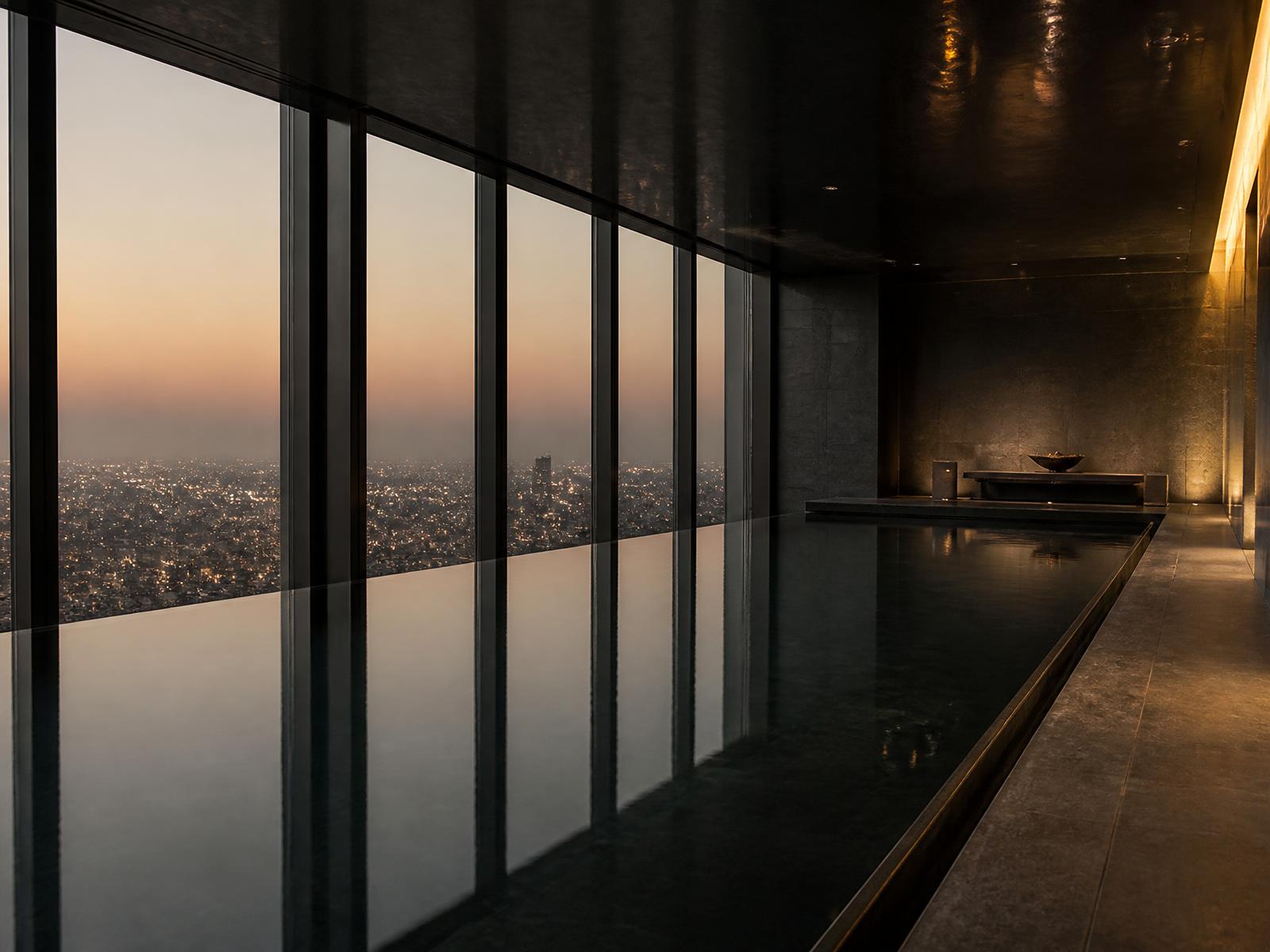 Long still indoor swimming pool in dark stone beside a floor-to-ceiling window overlooking Tokyo at dusk
