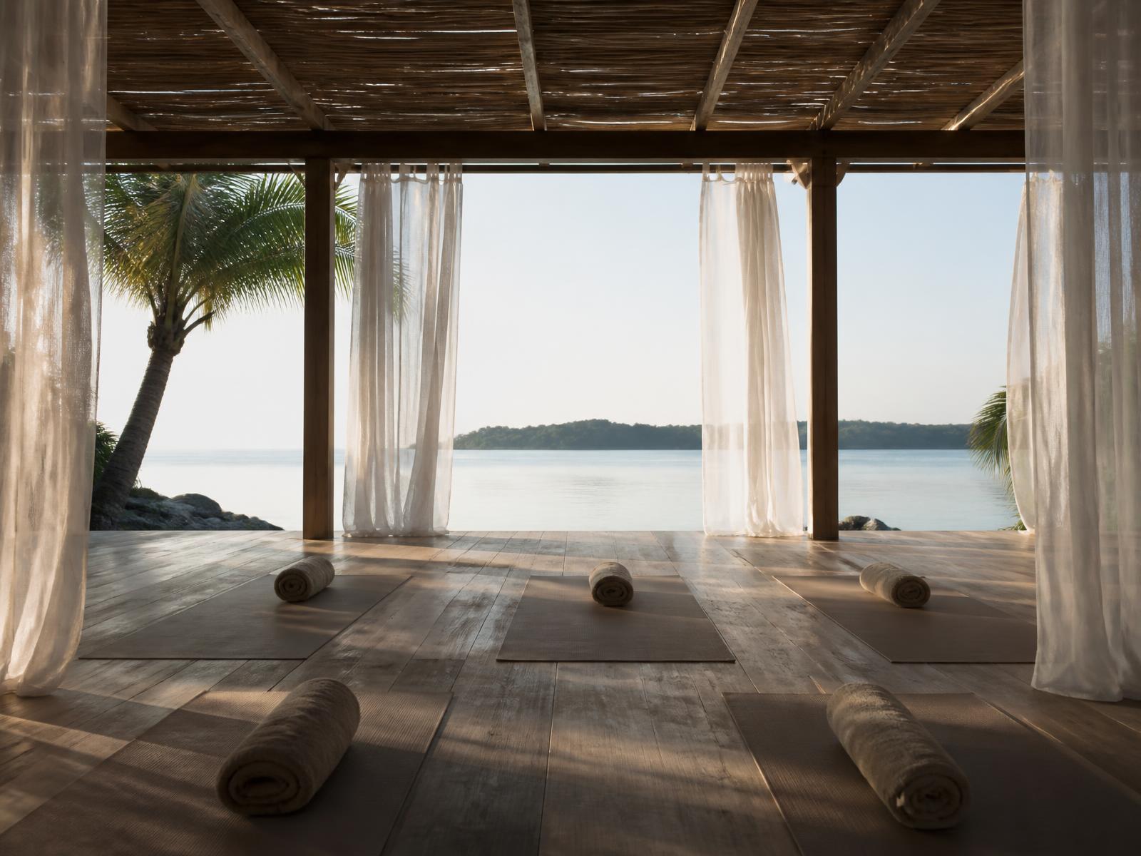 Open-air timber yoga pavilion facing calm turquoise water at dawn with rolled linen mats and sheer white curtains