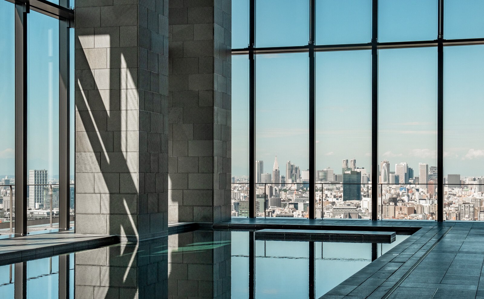 Ruhiges Innenschwimmbecken aus dunklem Stein an einer Glasfront mit weitem Blick über die Türme Tokios bei Tag