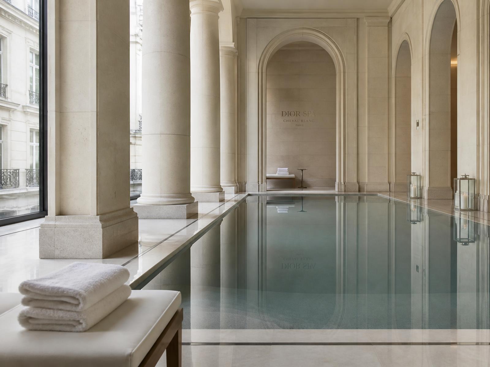 Still indoor swimming pool lined in pale limestone with columns, folded white towels and soft daylight