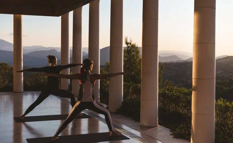 Zwei Personen beim Yoga auf einer Säulenterrasse in Amanzoe bei Sonnenaufgang, dahinter die Bergketten der Argolis im Dunst